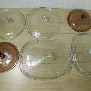May include: A collection of glass and brown-tinted glass cookware lids. The lids are round and oval, with a knob handle in the center. The lids are arranged on a light brown wooden surface.