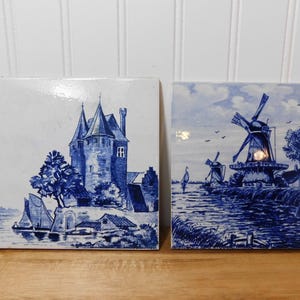 May include: Two square ceramic tiles with blue and white illustrations. One depicts a castle, the other windmills on a riverbank. The tiles are likely decorative and suitable for wall decor.