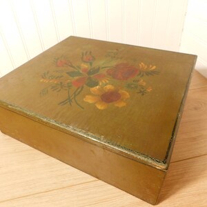 May include: A rectangular wooden box with a hinged lid. The lid is painted with a floral design in red, yellow, and green. The box is a muted olive green color and sits on a wooden surface.