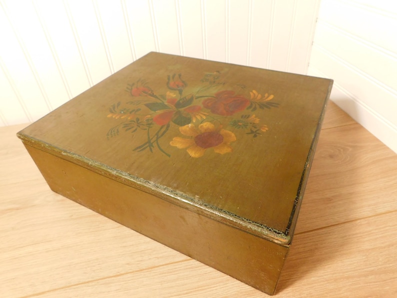 May include: A rectangular wooden box with a hinged lid. The lid is painted with a floral design in red, yellow, and green. The box is a muted olive green color and sits on a wooden surface.
