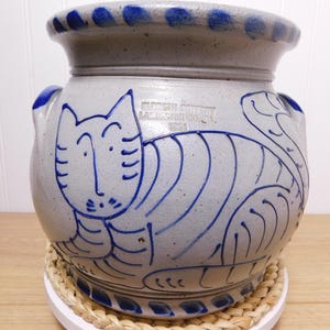 May include: A gray ceramic pot with a blue cat design. The pot has a wide, rounded shape with a flared rim and two small handles. The cat is depicted in blue lines, and the pot has a speckled texture. The pot is sitting on a woven mat.