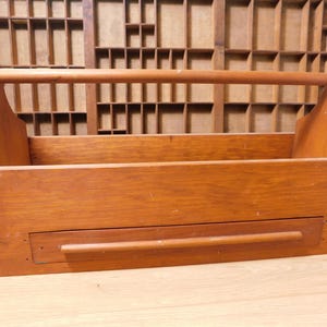 May include: A wooden tool tote with a handle, stained in a warm brown colour. The tote has a rectangular shape with a lower drawer and is designed for carrying tools or other items. The background features a vintage wooden printer's tray.