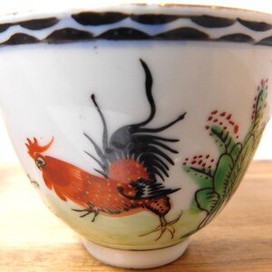 May include: White porcelain teacup with a hand-painted rooster design. The rooster is depicted in shades of red, orange, and black, with green and red accents. The rim features a black and blue wave pattern. A decorative piece for tea or coffee.