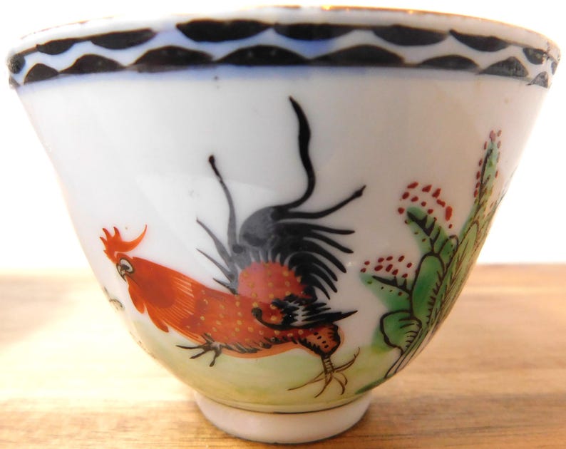 May include: White porcelain teacup with a hand-painted rooster design. The rooster is depicted in shades of red, orange, and black, with green and red accents. The rim features a black and blue wave pattern. A decorative piece for tea or coffee.