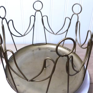 May include: A vintage brass-toned metal centerpiece. The design features a circular base with stylized figures holding hands around the rim. The figures have circular heads and curved arms, creating an open aesthetic. The piece sits on a white surface.