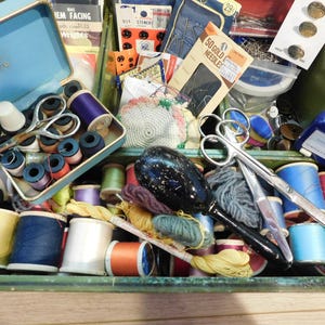 May include: A vintage sewing kit with a variety of supplies. The kit includes spools of thread in various colors, scissors, needles, thimbles, and other sewing notions. The kit is housed in a green wooden box with a blue metal tray.