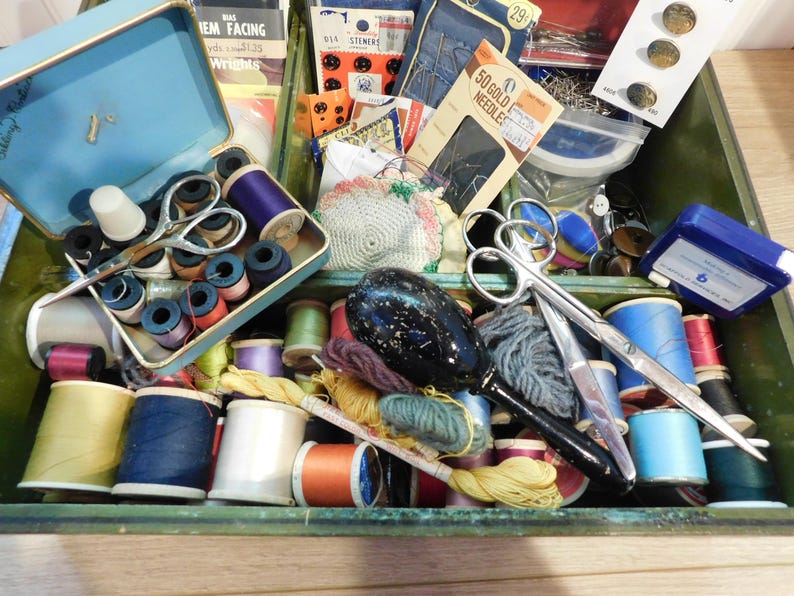 May include: A vintage sewing kit with a variety of supplies. The kit includes spools of thread in various colors, scissors, needles, thimbles, and other sewing notions. The kit is housed in a green wooden box with a blue metal tray.