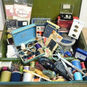 May include: Open green vintage sewing box filled with spools of thread in various colors, scissors, needles, buttons, and other sewing notions. A small blue sewing kit is open, revealing spools of thread and a thimble.