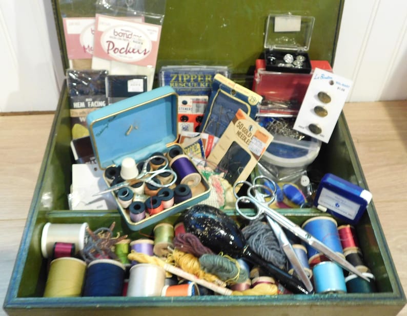 May include: Open green vintage sewing box filled with spools of thread in various colors, scissors, needles, buttons, and other sewing notions. A small blue sewing kit is open, revealing spools of thread and a thimble.