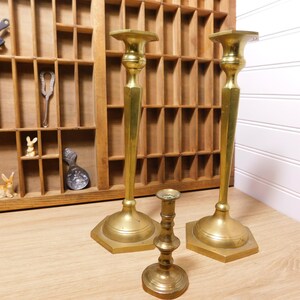 May include: Three brass candle holders of varying heights. Two are tall with hexagonal bases, and one is shorter with a round base. A wooden printer's tray with small decorative items is in the background.