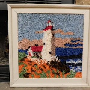 May include: A framed, textured artwork depicting a lighthouse scene. The lighthouse, with a red roof and white tower, stands on a rocky, colorful terrain. The sky is blue, and the sea is a deep blue. The artwork is in a white frame.