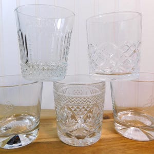 Pode incluir: Cinco copos transparentes com padr&otilde;es gravados decorativos. Os copos apresentam desenhos de diamantes e linhas verticais. Alguns t&ecirc;m uma borda inclinada e uma base decorativa. Os copos est&atilde;o dispostos sobre uma superf&iacute;cie de madeira.