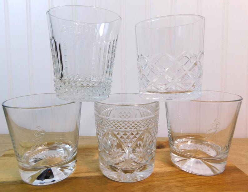 Pode incluir: Cinco copos transparentes com padr&otilde;es gravados decorativos. Os copos apresentam desenhos de diamantes e linhas verticais. Alguns t&ecirc;m uma borda inclinada e uma base decorativa. Os copos est&atilde;o dispostos sobre uma superf&iacute;cie de madeira.