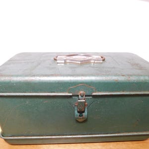 May include: A vintage, rectangular metal box in a faded green color. The box has a hinged lid with a metal clasp and a decorative handle. The metal shows signs of wear and age, with some rust visible.