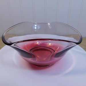 May include: A clear glass bowl with a deep red interior, placed on a white plate. The bowl has a distinctive, curved shape with a dark rim, contrasting with the red centre. The design is simple but elegant.
