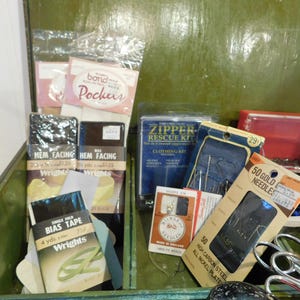 May include: An open green wooden sewing box filled with various sewing supplies. Items include packages of bias tape, hem facing, needles, a zipper repair kit, and scissors. The box contains a variety of sewing notions.