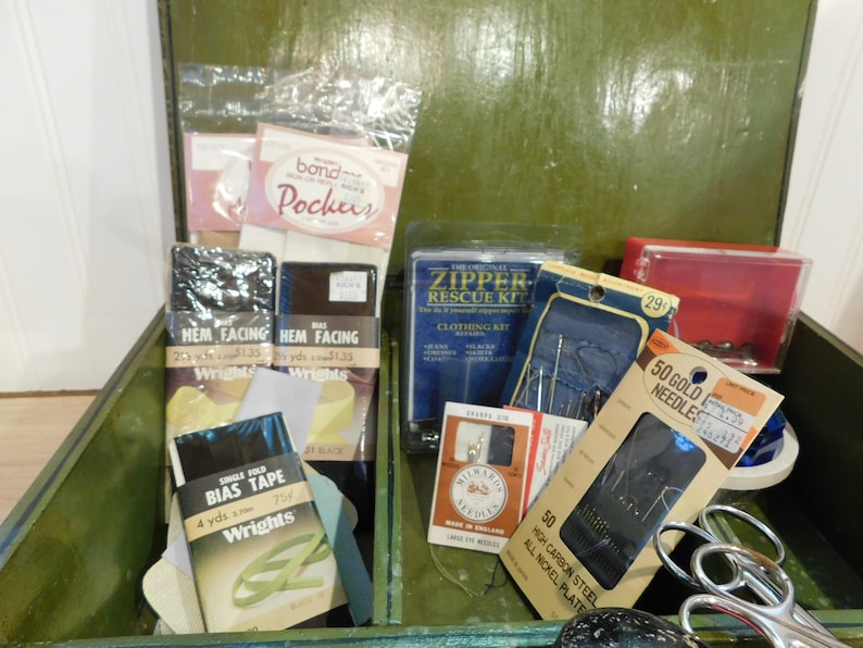May include: An open green wooden sewing box filled with various sewing supplies. Items include packages of bias tape, hem facing, needles, a zipper repair kit, and scissors. The box contains a variety of sewing notions.