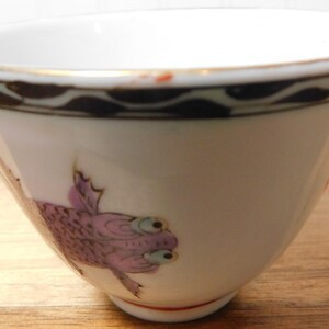 May include: White ceramic bowl with a gold rim and a decorative border. The bowl features a purple fish design on the side. The bowl is set on a wooden surface. This bowl is suitable for serving soups, cereals, or desserts.