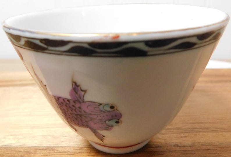 May include: White ceramic bowl with a gold rim and a decorative border. The bowl features a purple fish design on the side. The bowl is set on a wooden surface. This bowl is suitable for serving soups, cereals, or desserts.