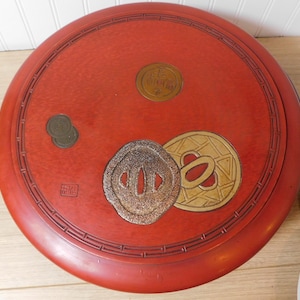 May include: A round, red wooden container with decorative gold and bronze accents. The lid features several circular and geometric designs, including a coin-like emblem and a stylized floral pattern. The container has a dark, intricate border.