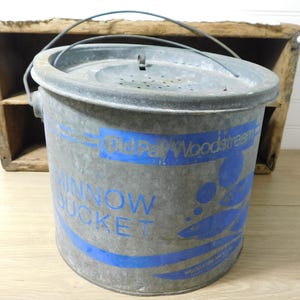May include: A galvanized metal minnow bucket with a wire handle and a perforated lid. The exterior features blue printed text reading Old Pal Woodstream Minnow Bucket and a graphic of fish. The bucket has a cylindrical shape and a weathered finish.