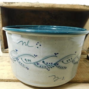 May include: A round ceramic bowl with a speckled off-white glaze and a dark teal rim. The exterior features a hand-painted horizontal vine pattern in dark teal and blue, consisting of curved lines, small dots, and leaf-like markings.