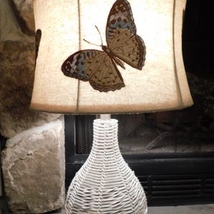 May include: A decorative table lamp with a white woven base and a beige lampshade. The lampshade features a large embroidered butterfly with brown and blue details. The lamp is plugged in and ready to illuminate a room.