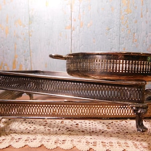 May include: Two rectangular silver-plated serving trays with ornate openwork gallery sides and decorative claw feet, stacked one atop the other. A round silver-plated serving dish with a matching pierced gallery rim rests on the top tray.
