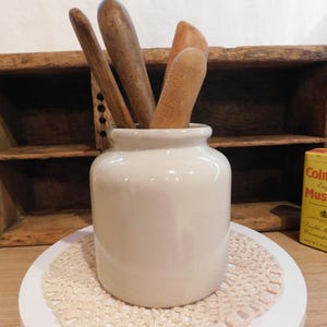 May include: A white ceramic crock containing three wooden kitchen utensils, positioned next to a yellow Colman's English Mustard tin. The crock has a smooth, cylindrical body with a rounded rim, and the wooden tools show natural grain textures.
