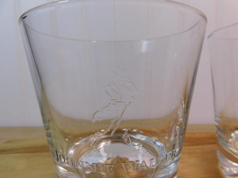 Pode incluir: Copo transparente e c&ocirc;nico com o logotipo Johnnie Walker gravado na frente. O logotipo apresenta uma figura a andar. As palavras "JOHNNIE WALKER" est&atilde;o gravadas na base do copo. O copo est&aacute; sobre uma superf&iacute;cie de madeira.
