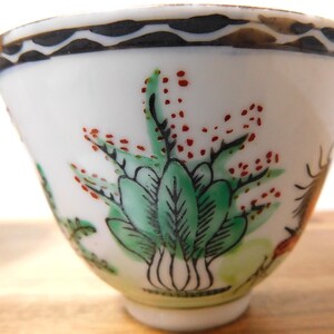 May include: A small, white porcelain teacup with hand-painted botanical designs. The cup features green leaves, red berries, and a black and brown decorative rim. The cup has a flared shape and a small base.