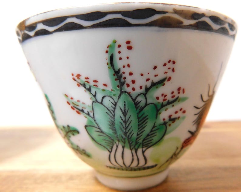 May include: A small, white porcelain teacup with hand-painted botanical designs. The cup features green leaves, red berries, and a black and brown decorative rim. The cup has a flared shape and a small base.