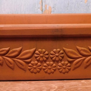 May include: A rectangular terracotta planter featuring a raised relief design on the front. The decorative pattern consists of symmetrical leafy branches extending from the sides toward a central cluster of five small, circular flower motifs.