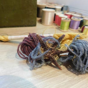 May include: A starfish rests amidst skeins of yarn in shades of purple, blue, and yellow. Spools of thread in various colors are in the background. The scene suggests a craft or hobby setting, possibly related to knitting or other textile arts.