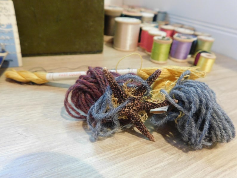 May include: A starfish rests amidst skeins of yarn in shades of purple, blue, and yellow. Spools of thread in various colors are in the background. The scene suggests a craft or hobby setting, possibly related to knitting or other textile arts.