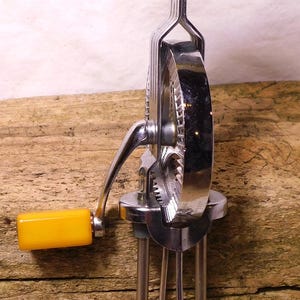 May include: A vintage hand-cranked egg beater with a chrome metal frame, two wire beaters, and a yellow wooden top handle showing signs of wear. A smaller yellow plastic handle is attached to the side crank, which rotates a central gear.