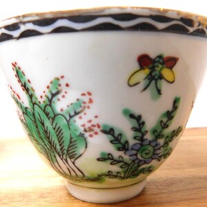 May include: A small, white porcelain teacup with hand-painted floral and insect designs. The cup features green foliage with red accents, a yellow and red insect, and a black and white wavy border around the rim. The cup sits on a small, white base.