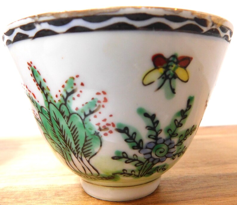 May include: A small, white porcelain teacup with hand-painted floral and insect designs. The cup features green foliage with red accents, a yellow and red insect, and a black and white wavy border around the rim. The cup sits on a small, white base.
