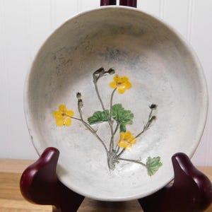 May include: A handmade ceramic bowl with a floral design. The bowl is off-white with a speckled texture and features a painted branch with yellow flowers and green leaves. The bowl is displayed on a wooden stand.