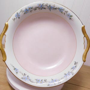 May include: A round, vintage serving bowl with a light pink center and white rim. The rim is decorated with a delicate floral pattern in blue and green. Gold-colored handles are on either side of the bowl.