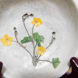 May include: A white ceramic bowl with a floral design. The bowl features a hand-painted image of yellow flowers, green leaves, and brown stems. The bowl is round and has a slightly textured surface. The bowl is resting on a dark brown stand.