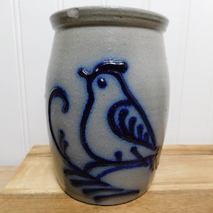 May include: A grey ceramic jar featuring a blue bird design. The bird is depicted on a branch, and the jar has a slightly textured surface. The rim of the jar is a darker grey colour.