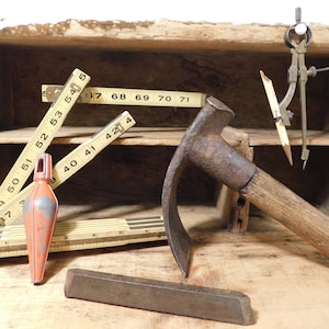May include: A collection of vintage tools including a wooden folding ruler with visible inch markings, a metal plumb bob with orange paint, a rusted metal adze with a wooden handle, a metal compass with a pencil, and a rectangular metal bar.
