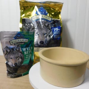 May include: Image of dog food and treats with a ceramic bowl. The food packaging features a wolf image and the words "Wilderness" and "Wild Bits". The bowl is beige and sits on a white surface.