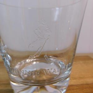 Pode incluir: Copo de vidro transparente com base espessa, com a imagem gravada de um homem a andar e as palavras "JOHNNIE WALKER". O copo tem uma forma alargada, ideal para bebidas espirituosas.