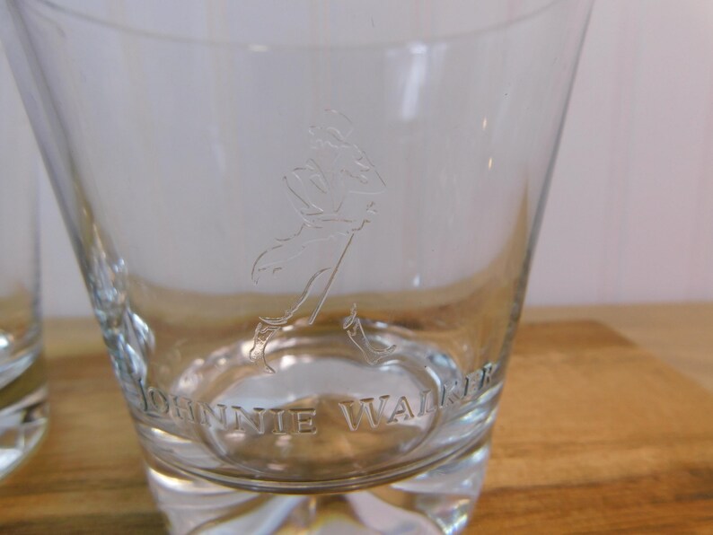 Pode incluir: Copo de vidro transparente com base espessa, com a imagem gravada de um homem a andar e as palavras "JOHNNIE WALKER". O copo tem uma forma alargada, ideal para bebidas espirituosas.