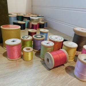 May include: A collection of vintage wooden spools filled with colorful threads. The spools vary in size and are filled with threads in shades of pink, yellow, green, purple, orange, and brown. Some spools have decorative paper labels.