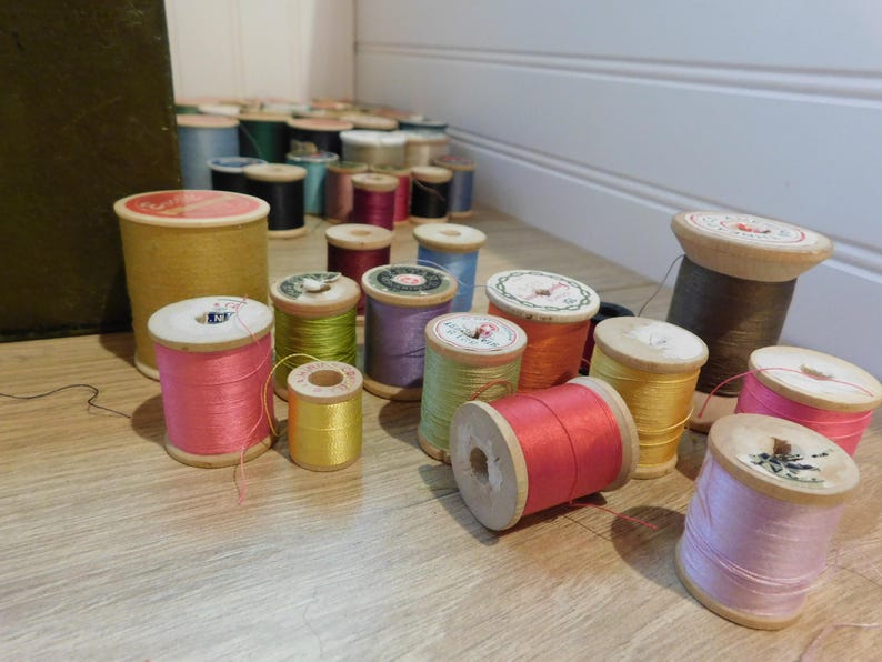May include: A collection of vintage wooden spools filled with colorful threads. The spools vary in size and are filled with threads in shades of pink, yellow, green, purple, orange, and brown. Some spools have decorative paper labels.