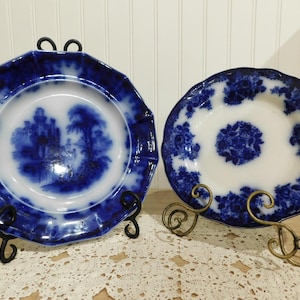 May include: Two antique blue and white decorative plates on display. One plate is octagonal with a landscape scene, the other is round with a floral design. Both are on decorative metal stands.