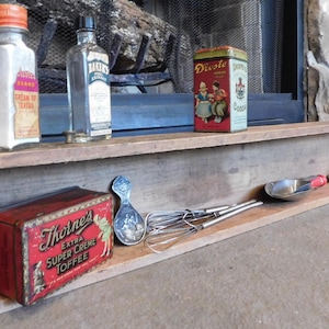 May include: A wooden shelf displays various vintage kitchen items. There are glass bottles, a red tin of cocoa, and a red tin of toffee. A metal whisk, a scoop, and a decorative spoon are also present. The shelf is set against a dark background.